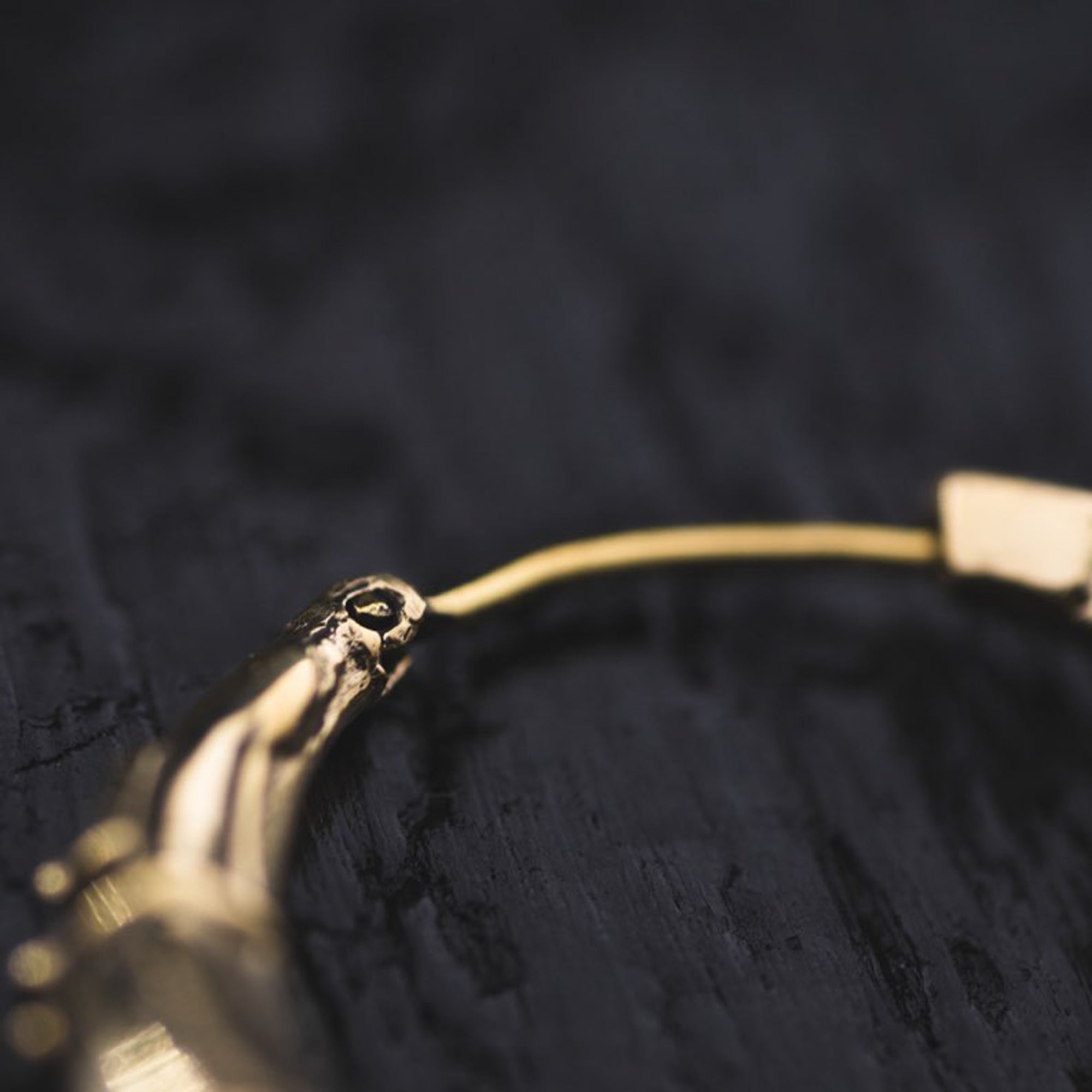 Close-up image of a partially visible PROMETHEUS Small Cyborg Hoop Earring in Gold by Kaya Movement on a dark surface. The earring showcases intricate detailing and a smooth, polished finish. The dark background contrasts with the bright gold, highlighting the earring's elegant design, reminiscent of post-apocalyptic body jewelry.